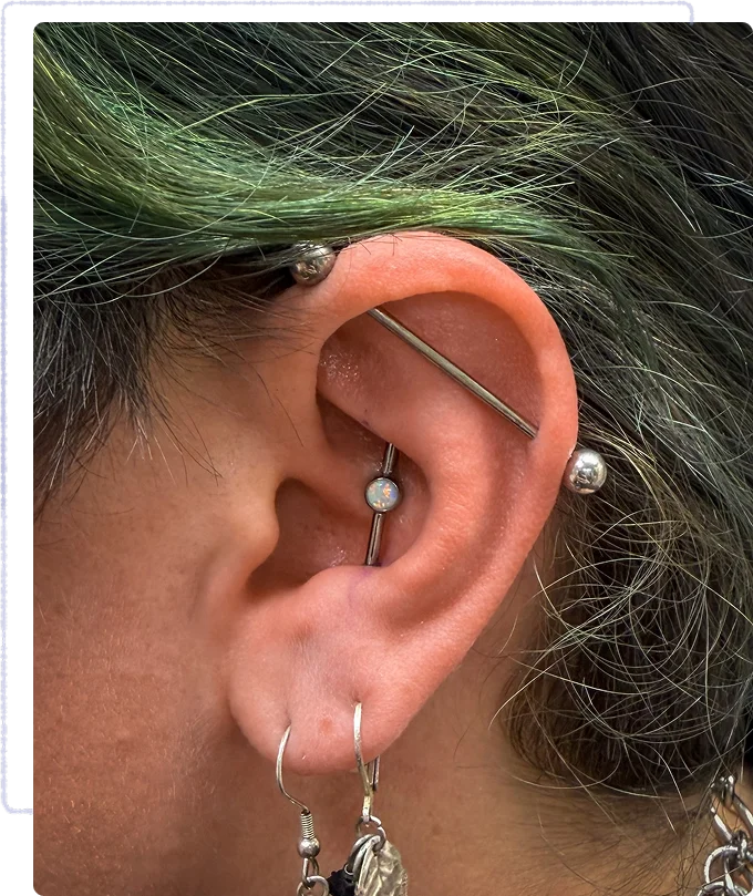Close-up of an ear with multiple piercings, including an industrial barbell, a conch stud, and two lobe earrings. Hair near the ear is dyed green.