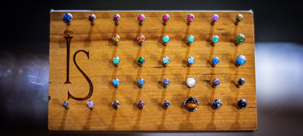 A wooden display board shows 36 pairs of colorful stud earrings, arranged in rows, with a stylized "IS" logo engraved on the left side.