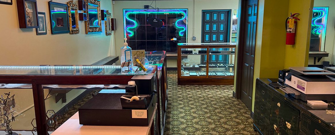 Interior of a jewelry or pawn shop with glass display cases, patterned carpet, neon signs in the window, and various items on display.