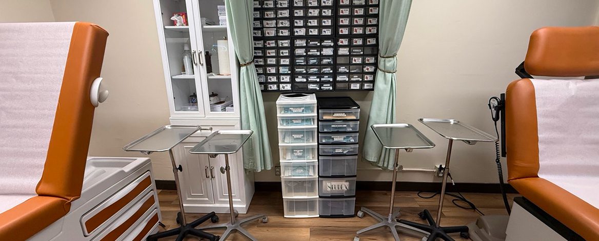 Medical exam room with two orange patient chairs, metal tray tables, storage drawers, cabinets, and a wall organizer with labeled compartments.