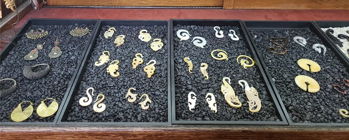 Four trays display various pairs of ornate earrings and ear gauges in different shapes and colors on a bed of black gravel.